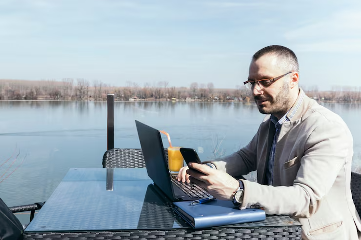 Man working on laptop and phone by the water, marine industry marketing